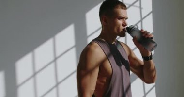 Atletik adam, güneşli bir stüdyoda egzersiz yaparken su içer. Sağlıklı yaşam tarzı seçimlerini örnek alır. Parlak Güneş Işığı Stüdyosunda Su İçen Atletik Genç Adam
