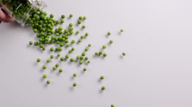 The mans hand rolls a jar full of green peas, which fills the surface. top view. Raw green peas in slow motion on white background.