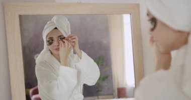 Bornozlu ve kafasında havlu olan genç bir kadın sağlıklı ciltli aynaya bakıp kahkahalarla kahkahalar atıyor ve göz torbalarını küçültüyor. Kozmetik ve yaş karşıtı estetik cerrahi.