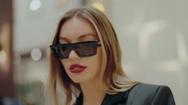 Kendine güvenen, şık güneş gözlüğü takan ve siyah ceketli bir kadın içeride poz veriyor, zarafet ve incelik yayıyor. Moda, güven ve modern tarz kavramları için mükemmel. Güneş Gözlüklü Kadın