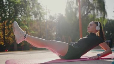 Beyaz kulaklıkla müzik dinlerken, parkta pembe bir minder üzerinde egzersiz yapan zinde bir genç kadın. Açık havada egzersiz yapan, güneşli bir günde müzikten zevk alan genç bir kadın.