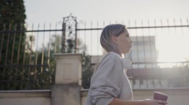 Genç bir kadın, sakin bir şehir ortamında sabah güneşini kucaklayarak kahvesini yudumlayarak huzur ve yeni bir başlangıç hissi veriyor. Sunlit City 'de Sabah Kahvesi İçen Genç Kadın