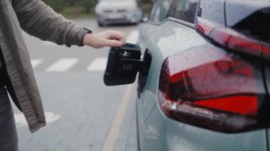 Yağmurlu bir günde şarj cihazını elektrikli araca takan genç bir adam sürdürülebilir ulaşımı gösteriyor. Şarj istasyonunda yağmurlu bir günde adam elektrikli arabayı şarj ediyor.