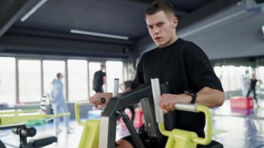 İyi donanımlı, parlak bir spor salonunda arka planda diğer insanlarla birlikte bacak pres makinesi kullanan odaklanmış genç bir erkek atlet. Modern Spor Salonu 'nda Bacak Baskı Makinesi ile Çalışan Genç Adam