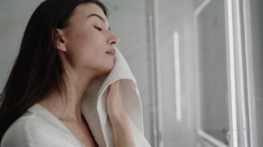 Banyoda yüzünü beyaz bir havluyla kurulayan genç bir kadın. Cilt bakımını tamamladıktan sonra rahatlamış görünüyor. Cilt rutininden sonra Banyoda Havluyla Kuruyan Kadın