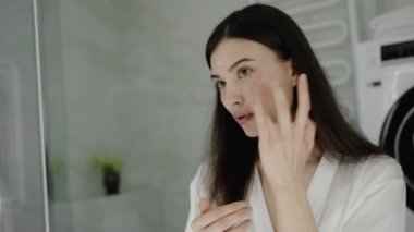 Modern bir banyo ortamında beyaz bornozlu genç bir kadın yüzüne cilt kremi sürüyor. Günlük kişisel bakım ve güzellik anlayışı. Sağlıklı Parlak Cilt İçin Cilt Kremi Uygulayan Kadın