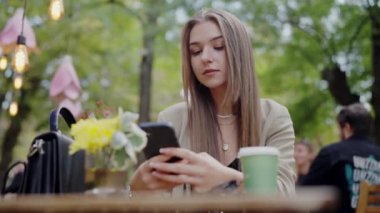 Genç bir kadın açık bir kafede oturuyor, akıllı telefonuna dalmış. Sahnede kentsel yeşillik ve yumuşak ışıklandırma yer alıyor. Açık Kafe Masasında Akıllı Telefon Kullanan Genç Kadın