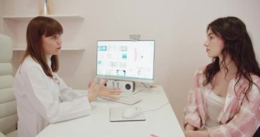 Kadın bir doktor, modern bir klinik ortamında görüntü sağlamak için bilgisayar ekranı kullanarak genç bir kadın hastayla sağlık detaylarını tartışıyor. Kadın Doktor Hastaya Sağlık Bilgilerini Açıklıyor