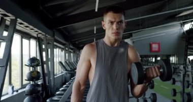 İyi donanımlı, modern bir spor salonunda dambılları kaldıran odaklanmış genç bir adam. Kendini spora adadığını gösteren yoğun bir ifade. Modern Spor Salonunda Ağırlık Kaldıran Genç Adam, Odaklanmış Spor