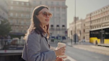 Güneş gözlüklü şık genç bir kadın güneşli bir şehir caddesinde sabah kahvesinin tadını çıkarıyor, modern şehir hayatı ve rahatlığı somutlaştırıyor. Şık Kadın Sunny City Sokak Sabahında Kahveyi Zevkle İçiyor