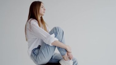 Gevşek beyaz gömlek ve mavi kot giyen sakin genç bir kadın, rahat bir poz oturarak düz bir arka plan karşı, yoğun ve düşünceli bir bakış yansıtır. Beyaz Gömlekli Serene Genç Kadın Pozisyonları