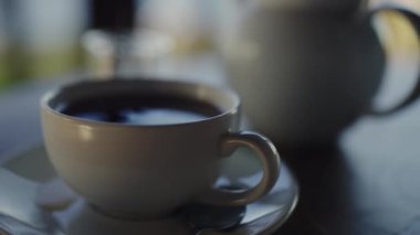 Buharı tüten bir çay fincanının yakın plan görüntüsü. Arka planda yumuşak odaklı çaydanlık, sakin bir ortamda yakalanmış. Sıcak bir ortamda Buharlı Çay Fincanının Yakın Çekimi