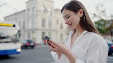 Genç bir kadın, arka planda yaklaşan bir otobüsle kalabalık bir şehir sokağında dururken akıllı telefonunu kullanarak nazikçe gülümsüyor. Modern kentsel yaşam tarzı ve bağlantı anlarını yakalar.