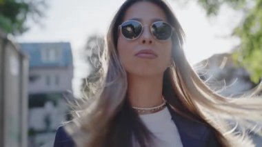 Parlak ve güneşli bir günde güneş gözlüğü takan modaya uygun genç bir kadının yakın plan portresi. Kendine güvenen ve modaya uygun. Güneş Gözlüklü Şık Genç Kadın Güneşli Şehir Gününün tadını çıkarıyor