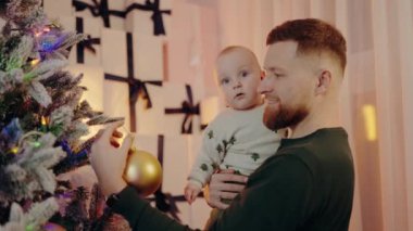Babam Noel ağacını süslerle süslerken kucağında bebek taşıyor. Paketlenmiş hediyelerle dolu şenlikli bir ortam. Tatil sezonunda aile bağını kutlamak.