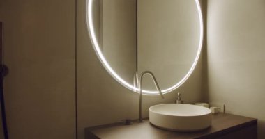 Geniş dairesel ışıklı ayna, parlak lavabo ve minimalist dekorun yer aldığı çağdaş bir banyo. Lüks iç tasarım kavramları ve mimari ilham için ideal..