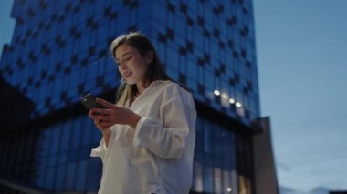 Akşamları akıllı telefonuyla şehir merkezindeki bir gökdelenin arka planına bakan genç bir kadın. Alacakaranlık 'ta Akıllı Telefon Kullanan Genç Kadın