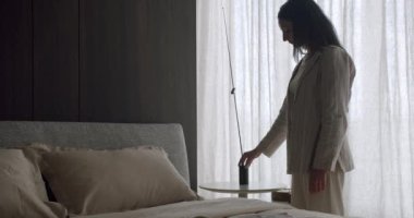 Minimalist bir yatak odasında lamba ayarlayan bir kadının yan profili. Sahne, modern tasarımı ve yumuşak ışıklandırmasıyla sakin ve sakin bir atmosfer yayıyor. Sakin Atmosferi olan Minimalist Yatak Odası