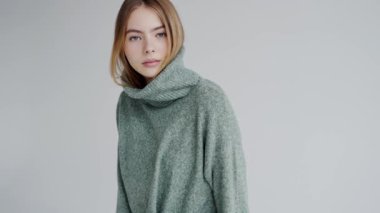 Sakin ve güzel bir genç kadın portresi, sarı saçlı, rahat yeşil balıkçı yaka kazak giymiş, tarafsız gri bir arka planda, sükuneti ve günlük tarzını tasvir eden.