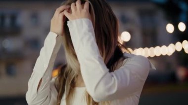 Beyaz elbiseli güzel bir kadının portresi gece açık havada duruyor, şehir ışıkları arka planda sıcak bir bokeh etkisi yaratıyor. Genç Güzel Kadın. Parlak Gözlü Genç Kadın Portresi.