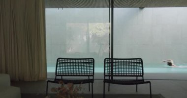 İç mekan, yüzen bir adam, modern banklar ve akarsu perdeleri olan şık bir kapalı havuz alanı gösteriyor. Modern Mobilya ve Perdelerle Kapalı Havuz Bölgesi