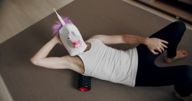 Tek boynuzlu at maskeli komik adam evde miyofasyal gevşeme için tekerlekli sandalyeyle egzersiz yapıyor. Evde komik bir spor salonu var. Kas ve fasya germe için köpük silindiri. Evde kendine masaj için aletler.