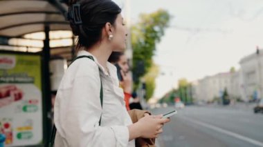Şehirdeki otobüs durağında bekleyen genç bir kadın, akıllı telefonuna dalmış. Kentsel yaşam tarzı, günlük ulaşım ve teknoloji kullanımı. Şehir Merkezinde Akıllı Telefon Kullanılırken Otobüs Durağında Bekleyen Kadın