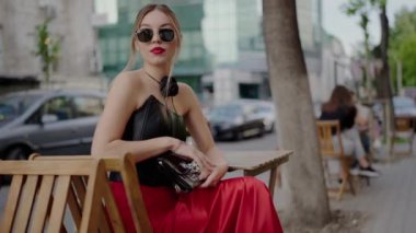 Güneş gözlüğü takan, deri bluzlu genç bir kadın şehirdeki açık bir kafede oturuyor ve çantasına bakıyor. Şehir hayatı ve sokak modası. Güneş Gözlüklü Moda Genç Kadın