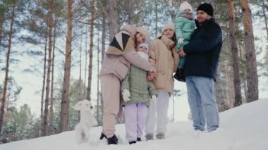Çocuklu bir aile ve uzun çam ağaçlarının arasında güzel bir kış günü geçiren küçük bir köpek. Hepsi kalın giysiler giyiyor ve neşeli ifadeler takınıyorlar. Mutlu Aile Karlı Günün tadını çıkarıyor