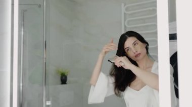 Banyoda saçını tarayan, maske takan bir kadın. Sahne, taze ve kendine özen gösteren bir atmosfer yayıyor. Banyoda Yama Maskeli ve Doğal Işıklı Kadın Saçları