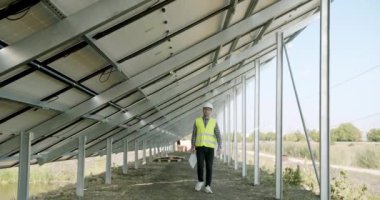 Erkek mühendis güneş panellerini inceliyor. Üniformalı bir yetişkin fotovoltaik panelleri kontrol ediyor ve güneş enerjisi istasyonuyla ilgili çalışırken günlüğe not alıyor.