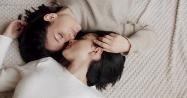 Low angle handheld shot of young lesbian couple smiling and touching hands gently while sitting on sofa against window in light living room at home. Lifestyle and relationships. LGBT concept