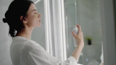 Siyah saçlı, beyaz cüppeli bir kadın banyo aynasının önüne saç spreyi sıkar. Görüntü, sakin bir günlük rutin, kişisel bakım ve modern bir ortam sunuyor..