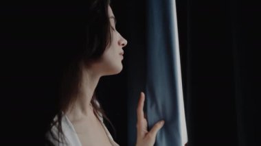 Pencerenin yanında durup, derin düşüncelere dalıp dışarı bakan bir kadın. Düşünceli ve içe dönük görünüyor, doğal ışık sahneyi aydınlatıyor. Pencereden Dışarı Bakan Kadın