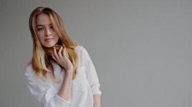 Uzun saçlı çekici bir genç kadın, minimalist bir düzende beyaz gömlek giyerken nazikçe gülümsüyor. Beyaz tişörtlü zarif genç kadın. Evinde usulca gülümsüyor.