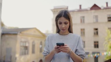 Sıradan kıyafetli odaklanmış genç bir kadın arka planda tarihi binalar ve sahneyi aydınlatan yumuşak bir güneş ışığıyla hareketli bir şehir ortamında akıllı telefonunu kullanır..