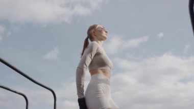 Bir bayan sporcunun dinamik görüntüsü açık hava fitness parkında yüksek atlama yaparak çeviklik ve gücü gösteriyor. Atletik vücutlu genç bir kadın fiziksel egzersiz yapıyor ve sokakta zıplıyor.