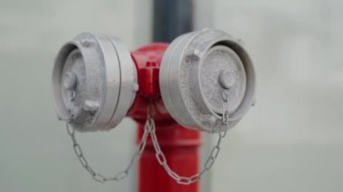 Metal kapaklı kırmızı bir yangın musluğunun ve bulanık bir şehir geçmişine bağlı zincirlerin detaylı görüntüsü, güvenlik ve belediye hizmetlerini vurguluyor. Kırmızı Yangın Yangın Yangın Musluğu Kapağı ve Zinciri