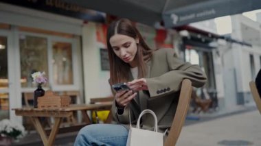 Telefonunu kullanmaya odaklanmış genç bir kadın arka planda hareketli şehir hayatı olan bir kafenin dışındaki küçük bir masada otururken. Açık Kafe Masasında Akıllı Telefon Kullanan Genç Kadın