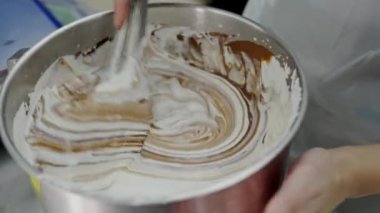 Bulanık bir elin, beyaz ve siyah çikolata kremiyle dolu bir çelik karıştırma kabını karıştırdığı, aşçılık becerilerini ve tatlı hazırlığını sembolize eden yakın plan görüntüsü. Çikolata kıvrımını kamçılamak Bir anlık bakış 