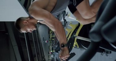 Genç bir atletik adam, modern bir spor salonundaki kardiyo makinesinde çalışırken akıllı yamayla performansını izliyor. Yoğun jimnastik yaparken Smartwatch kullanmaya odaklanmış genç adam. Dikey video.