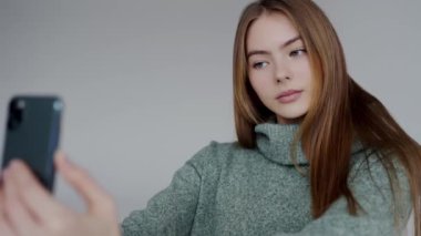 Genç kadın akıllı telefonuyla selfie seansının tadını çıkarıyor. Uzun saçlı gülümseyen genç bir kadın rahat bir kazakla selfie çekiyor. Bir anlık gençlik sevinci ve teknoloji kullanımını yakalar.