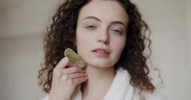 Yeşil bir Gua Sha taş kazıyıcısıyla yüz masajı yapan genç bir kadının portresi. Cilt kozmetolojisi kavramı. Doğal güzellik estetiği. Yüz bakımı ve güzellik ayinleri