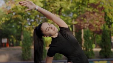 Genç bir kadın kollarını zarif bir şekilde sakin bir parkta uzatıyor, canlı yeşillikler arasında sağlığını ve sağlığını somutlaştırıyor. Park 'ta Kol Geren Genç Kadın, Dışarıda Egzersiz Yapıyor.