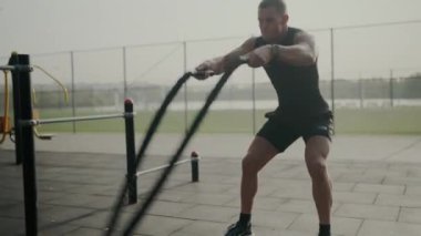 Siyah spor giyimli odaklanmış bir adam açık hava spor salonunda savaş halatlarıyla egzersiz yapıyor. Güç ve kararlılık gösteriyor. Açık Spor Salonu 'nda Savaş Halatlarıyla Çalışan Kararlı Adam