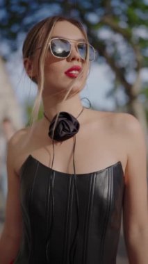 Güneş gözlüklü ve siyah elbiseli şık bir kadın güneşli bir günde şehir ortamında güven içinde poz veriyor. Görüntü zarafeti ve modern modayı yakalıyor. Güneş Gözlüklü Moda Kadını, Dikey.