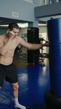 Çağdaş bir spor salonunda mavi bir çantayı yumruklayan kendini adamış genç bir adam antrenman sırasında gücünü ve dikkatini gösteriyor. Modern Spor Salonu 'na odaklanmış Genç Adam Kum Torbası. Dikey video.