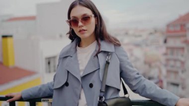 Moda güneş gözlüklü ve mavi trençkotlu şık genç bir kadın şehrin çatısında güven içinde duruyor, kentsel ve modaya uygun bir hava veriyor. Güneş gözlüklü şık kadın ve şehre tepeden bakan trençkot.