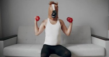 At maskeli komik bir adam evdeki beyaz koltukta ağırlık çalışması için egzersiz yapıyor. Evde komik bir spor salonu var. Komik Sağlık ve Spor Konsepti. Esprili esnemeler. Komik ev egzersizi.