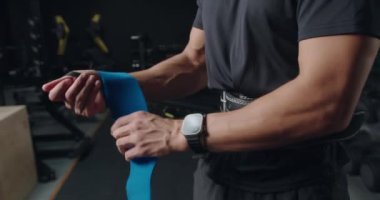 Modern bir spor salonunda mavi bilek desteği kullanarak spor yapmaya hazırlanan odaklanmış erkek sporcu, akıllı hareket gösteriyor. Atletik Adam Bileklerini Destek Bandıyla Spor Salonuna Sardı